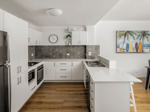 A modern kitchen with white cabinetry, a wooden floor, a clock, and a surfboard painting.