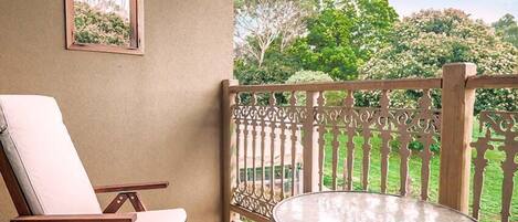 A balcony with a white cushioned chair and a small round table, overlooking a lush garden with trees and a fence.