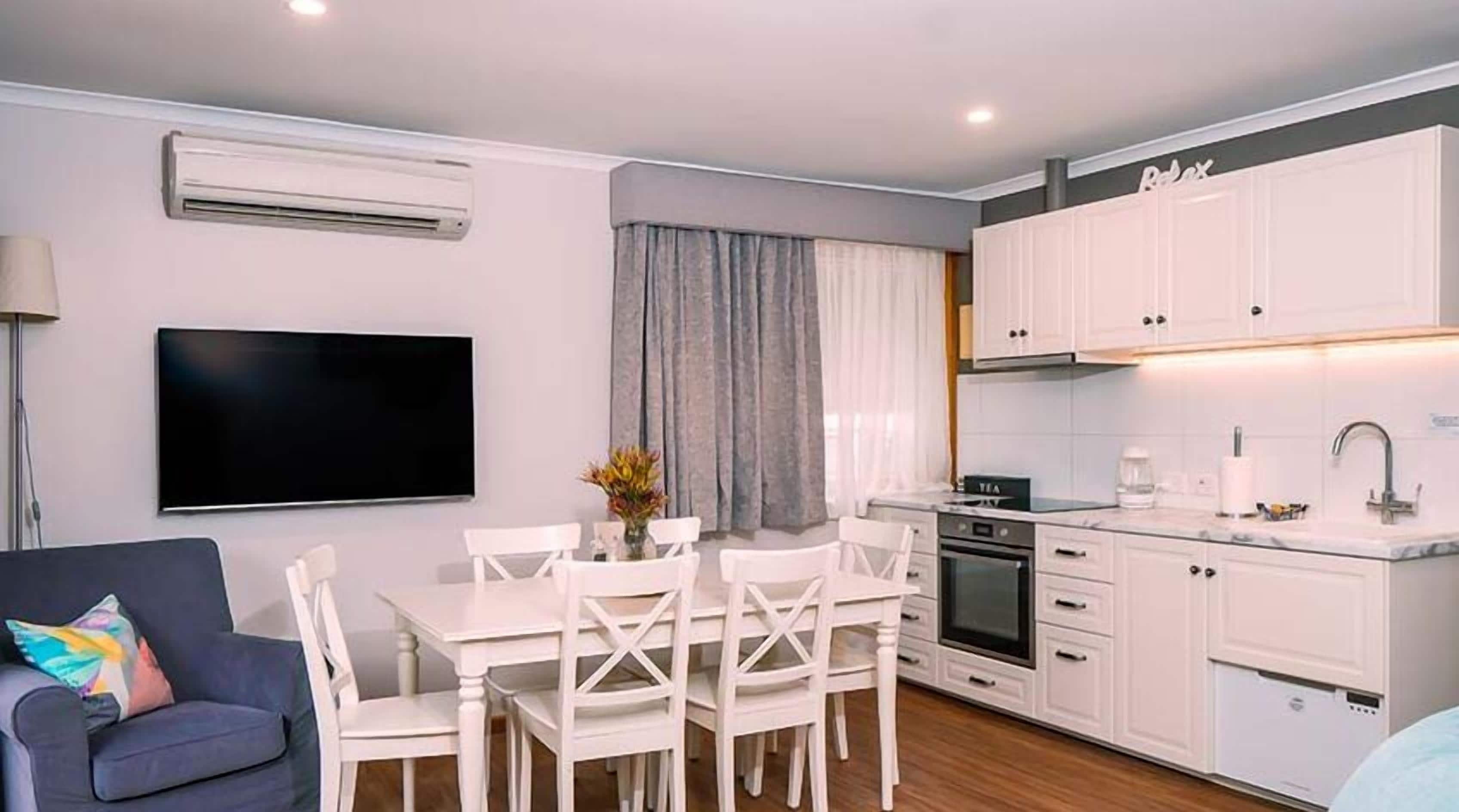 A modern kitchen with white cabinets, a built-in oven, and a sink. There is a dining area with a white table and chairs. A flat-screen TV is mounted on the wall.