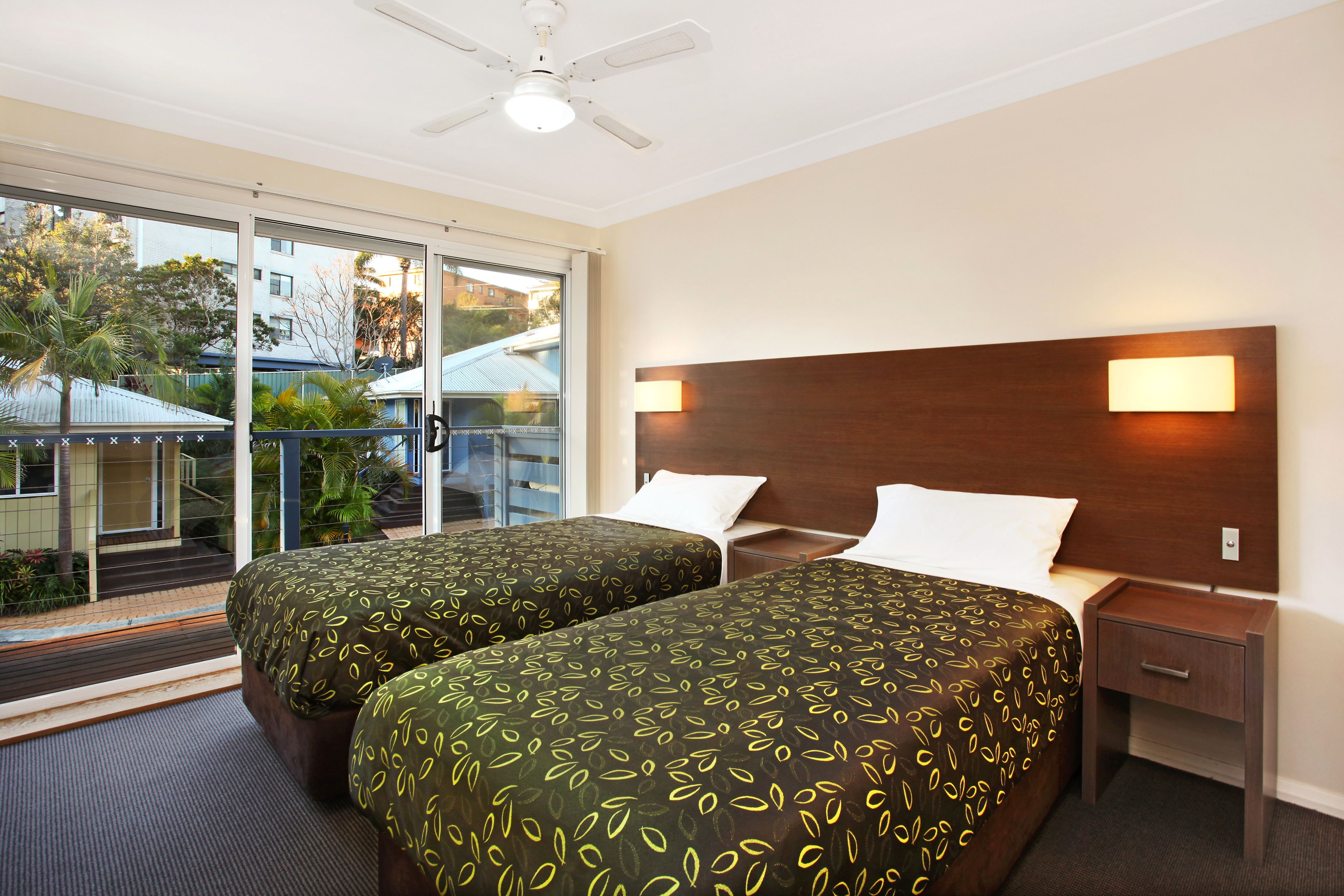 A hotel room with two beds, a nightstand, a ceiling fan, and a balcony with a view of buildings and trees.