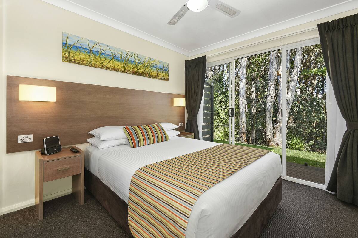 Una habitación de hotel con una cama grande, una mesita de noche con lámpara, un cuadro en la pared y vistas al exterior a través de una puerta corredera.