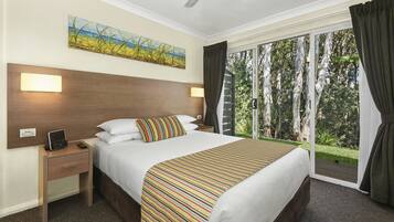 A hotel room with a large bed, a nightstand with a lamp, a painting on the wall, and a view of the outdoors through a sliding door.