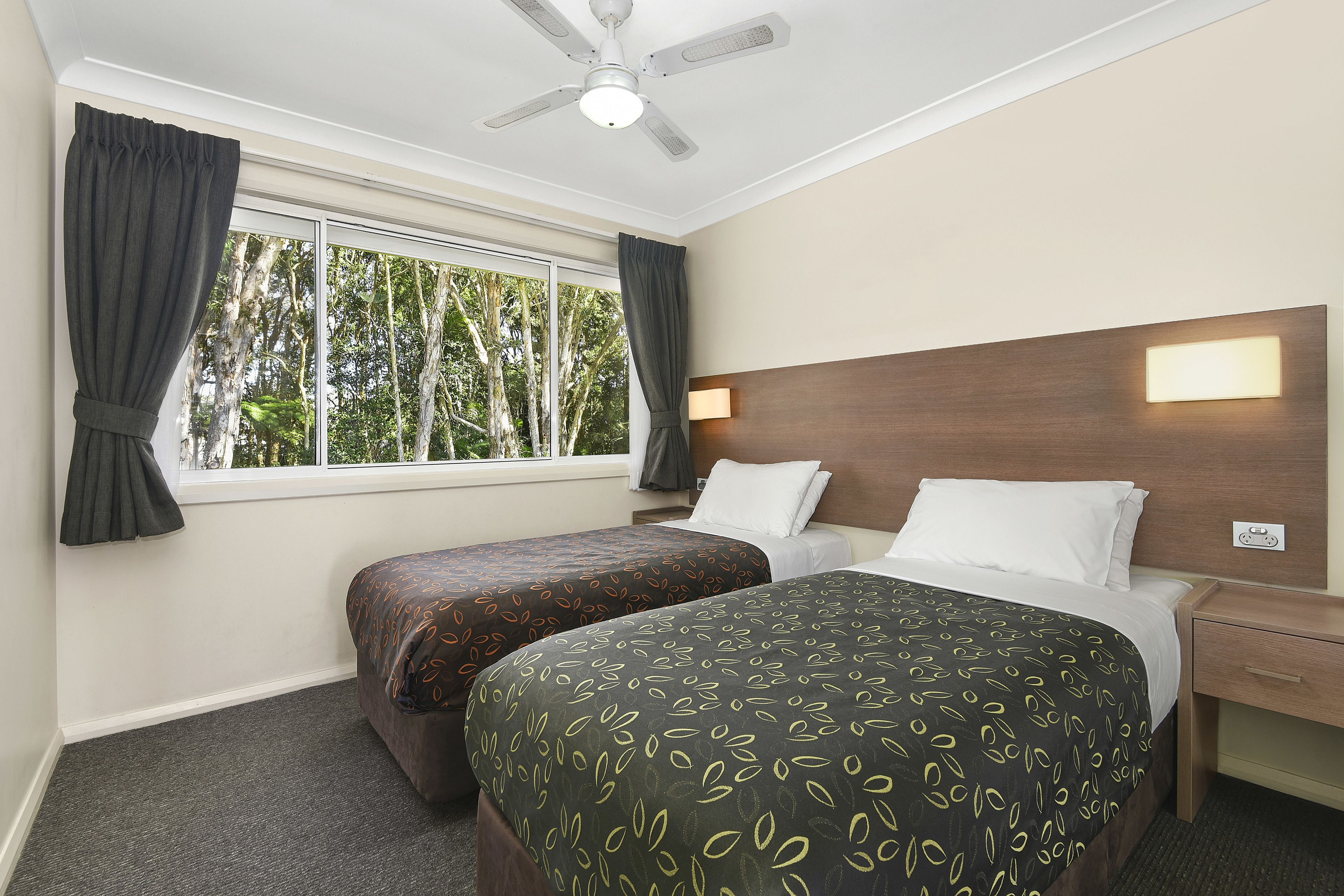 Habitación de hotel con dos camas, ventilador de techo, ventana con cortinas, mesita de noche y una lámpara fijada a la pared.