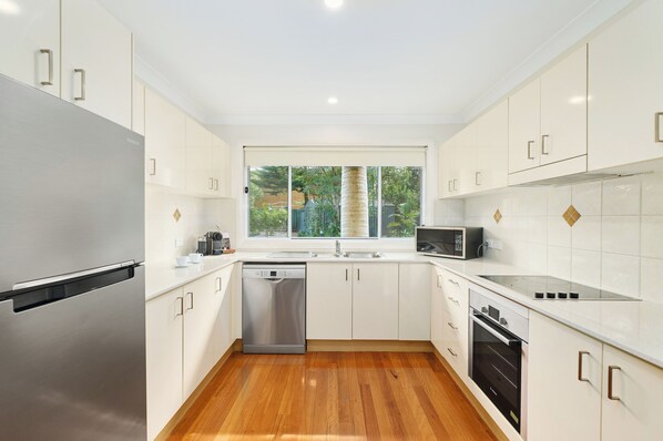 A modern kitchen with white cabinets, stainless steel appliances, and a wooden floor. - Flynns on Surf Beach Villas (Port Macquarie)