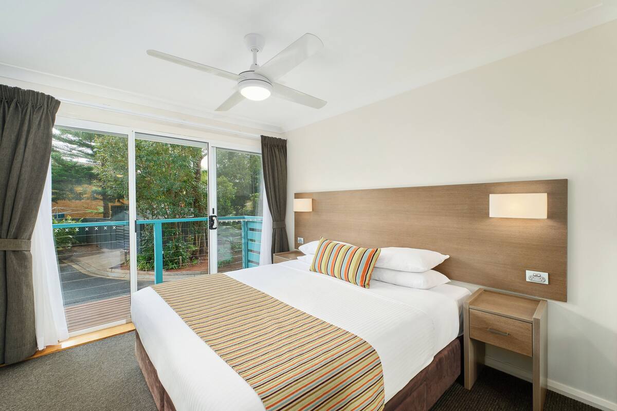 Habitación de hotel con una cama grande, ventilador de techo, mesita de noche y vistas a un balcón con barandilla.