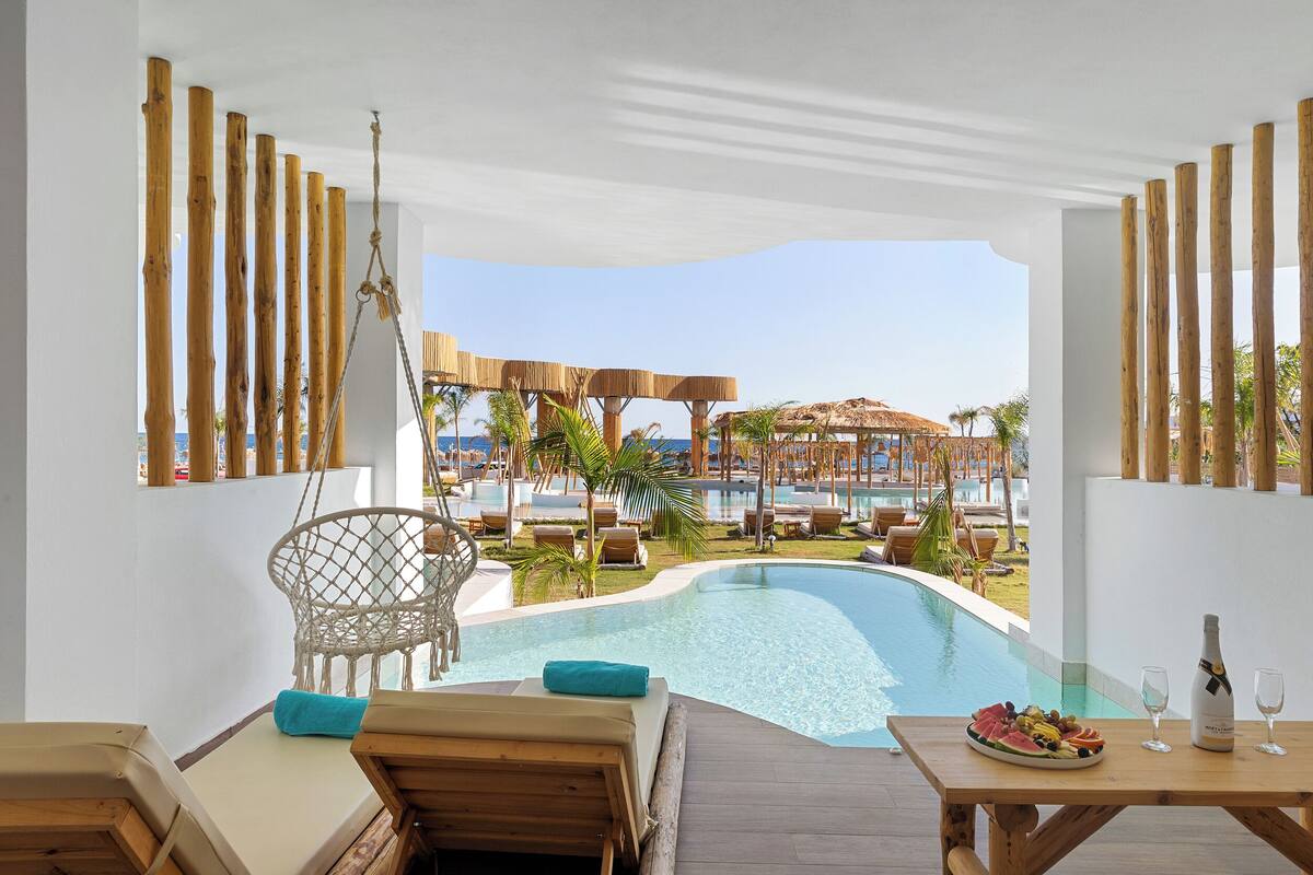 A poolside area with wooden furniture, a dining table with a plate of fruit and a bottle, and a view of a resort with palm trees and a pool.