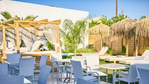 An outdoor seating area with white wicker chairs and tables, a wooden pergola with a white canopy, and thatched-roof structures.
