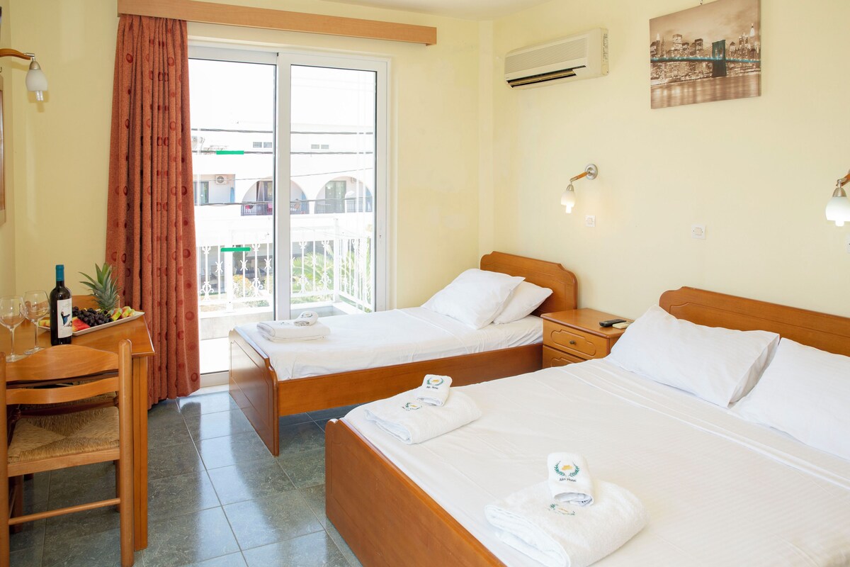 A hotel room with two beds, a dining table with a bottle and fruit, and a balcony with a city view.