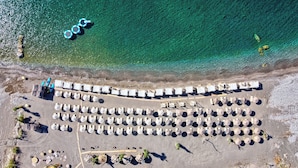 Vista aérea de un complejo turístico frente al mar, con hileras de sillas y sombrillas de playa, una estructura de madera y una vista despejada del océano.