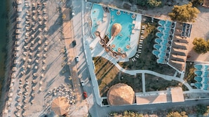 Vista aérea de un resort con un amplio espacio de piscina, numerosas tumbonas y una playa con sombrillas.