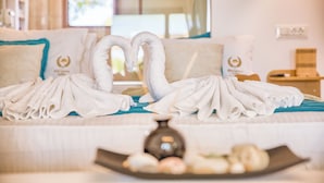 Habitación de hotel con cama, toallas dispuestas en forma de corazón, una bandeja con frutas y un jarrón.