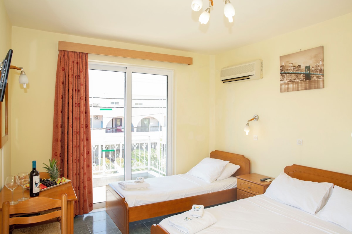 A hotel room with two beds, a balcony, a dining table with a bottle and glasses, and a cityscape view.