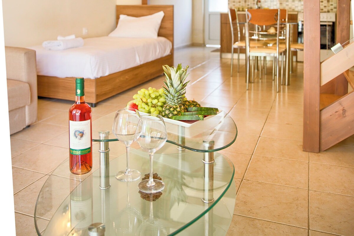 A hotel room with a bed, a glass table with a bottle and glasses, a fruit bowl, and a dining area with a table and chairs.