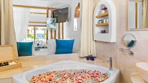 A modern bathroom with a large bathtub filled with rose petals, a wooden side table with toiletries, and a view of a balcony with blue cushions.