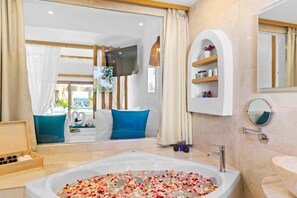Un baño moderno con una bañera grande llena de pétalos de rosa, una mesita de madera con artículos de aseo, y vistas a un balcón con cojines azules.