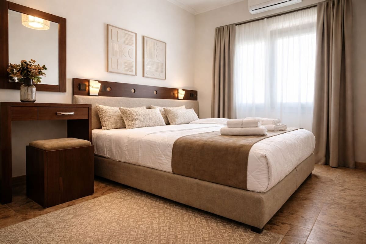 Una habitación de hotel con cama, mesita de noche, espejo, una obra de arte y una ventana con cortinas.