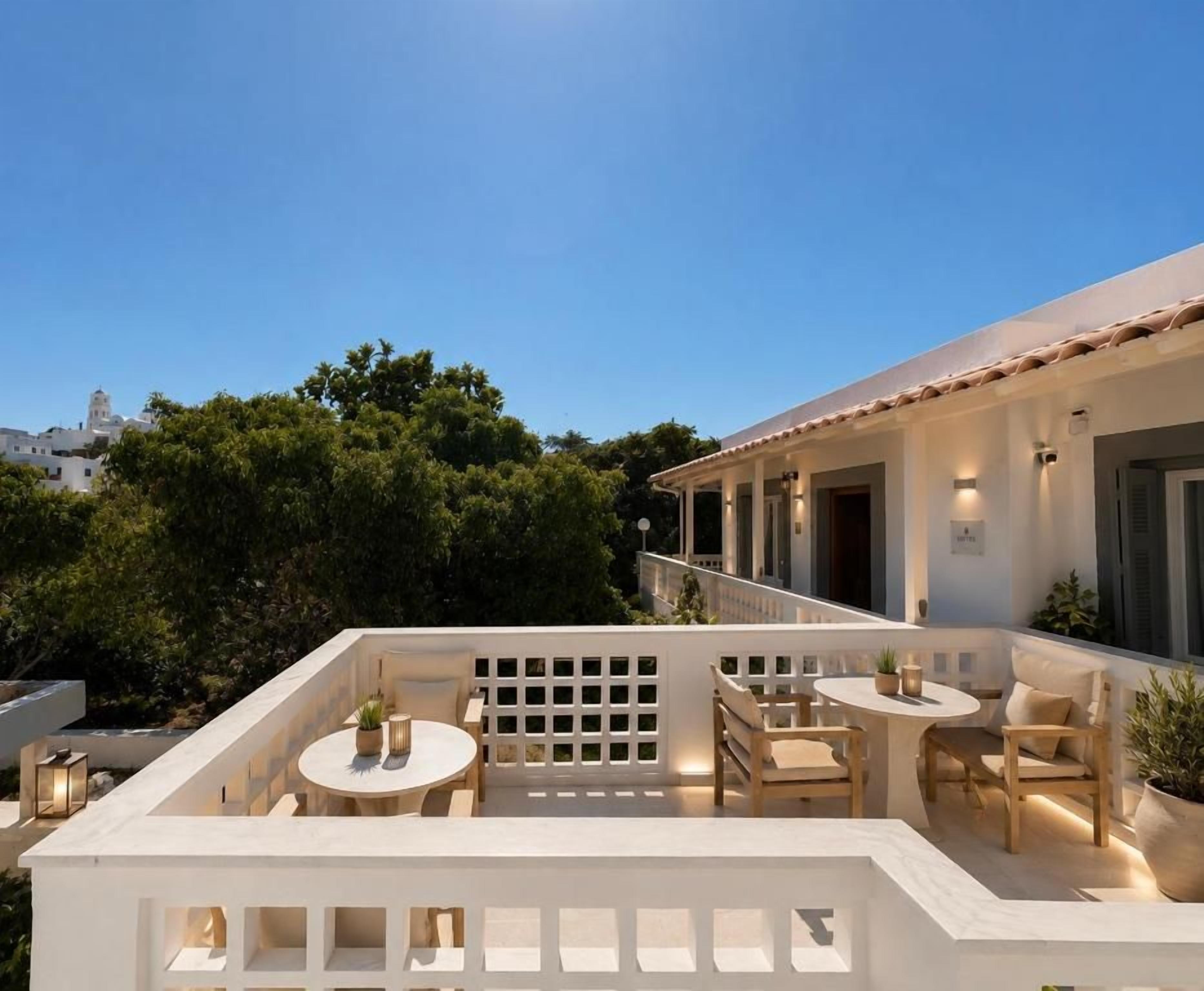 Un patio exterior con mesas y asientos, un edificio blanco con balcón y árboles al fondo.