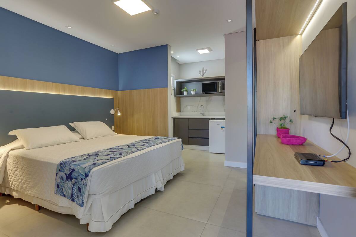 A modern bedroom with a bed, bedside table, and a small kitchenette in the background.