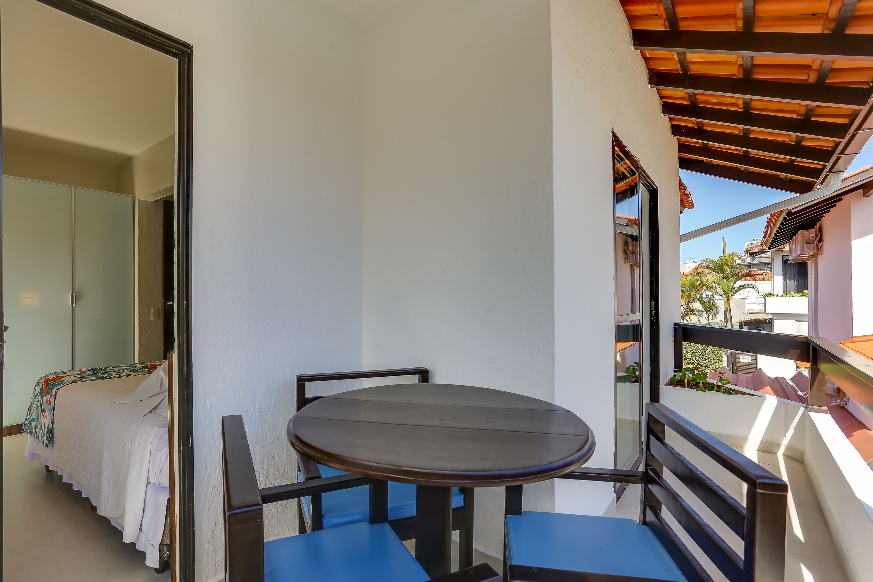 Varanda com mesa redonda e duas cadeiras, cama com colcha branca e vista para edifícios e palmeiras.