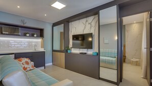 A modern living room with a sofa, a flat-screen TV, a kitchen area with a microwave, and a bathroom visible through an open door. - Hotel Sete Ilhas (Florianópolis)