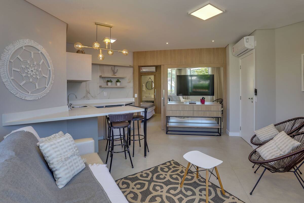A modern living room with a sofa, a coffee table, and a TV. There is a kitchen area with a sink and a dining table.