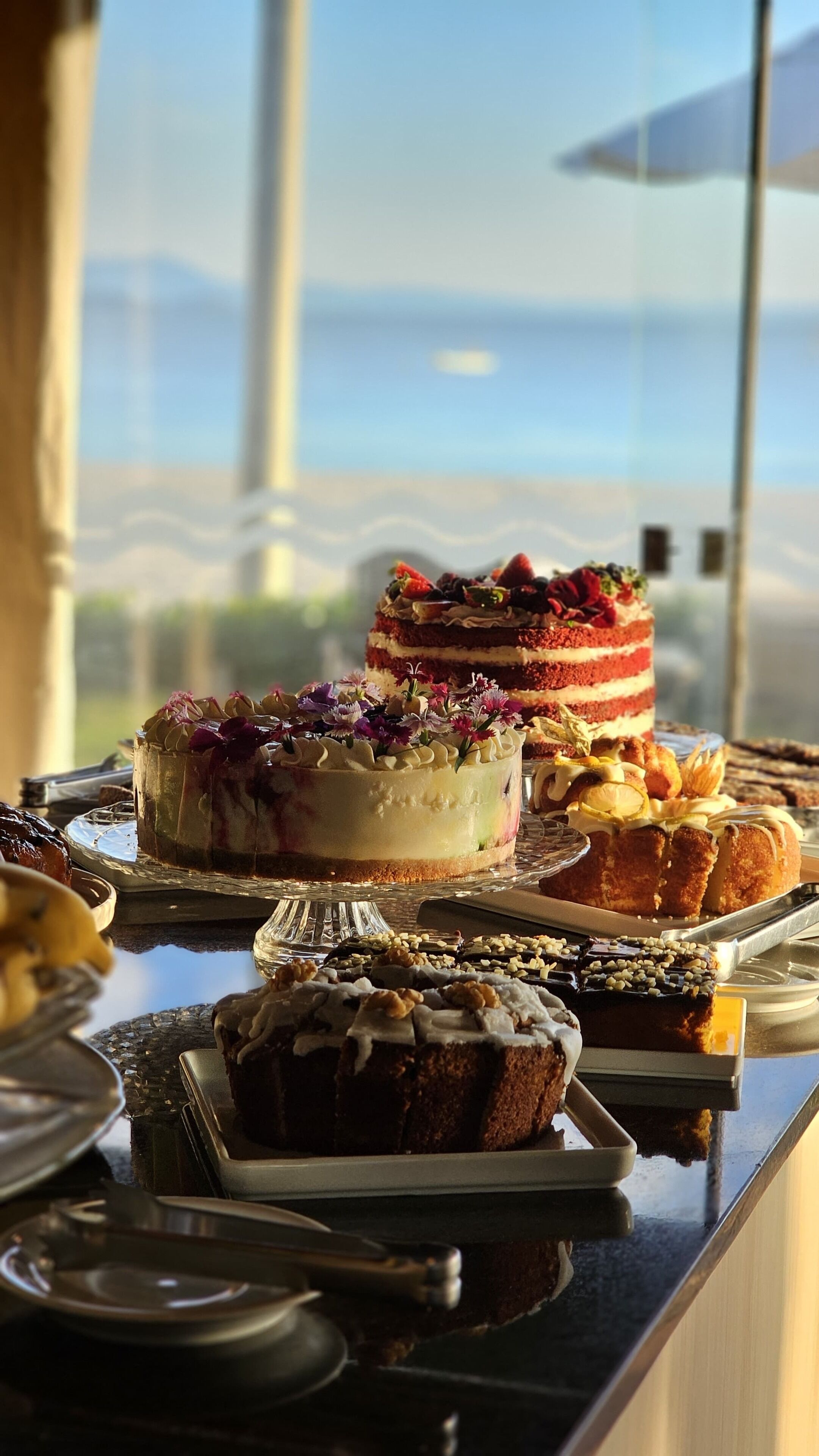 A dessert buffet with various cakes and pastries, arranged on a table with a view of the ocean in the background.