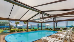 An indoor swimming pool with lounge chairs, a view of the sea, and a covered area with a metal roof. - Hotel Sete Ilhas (Florianópolis)
