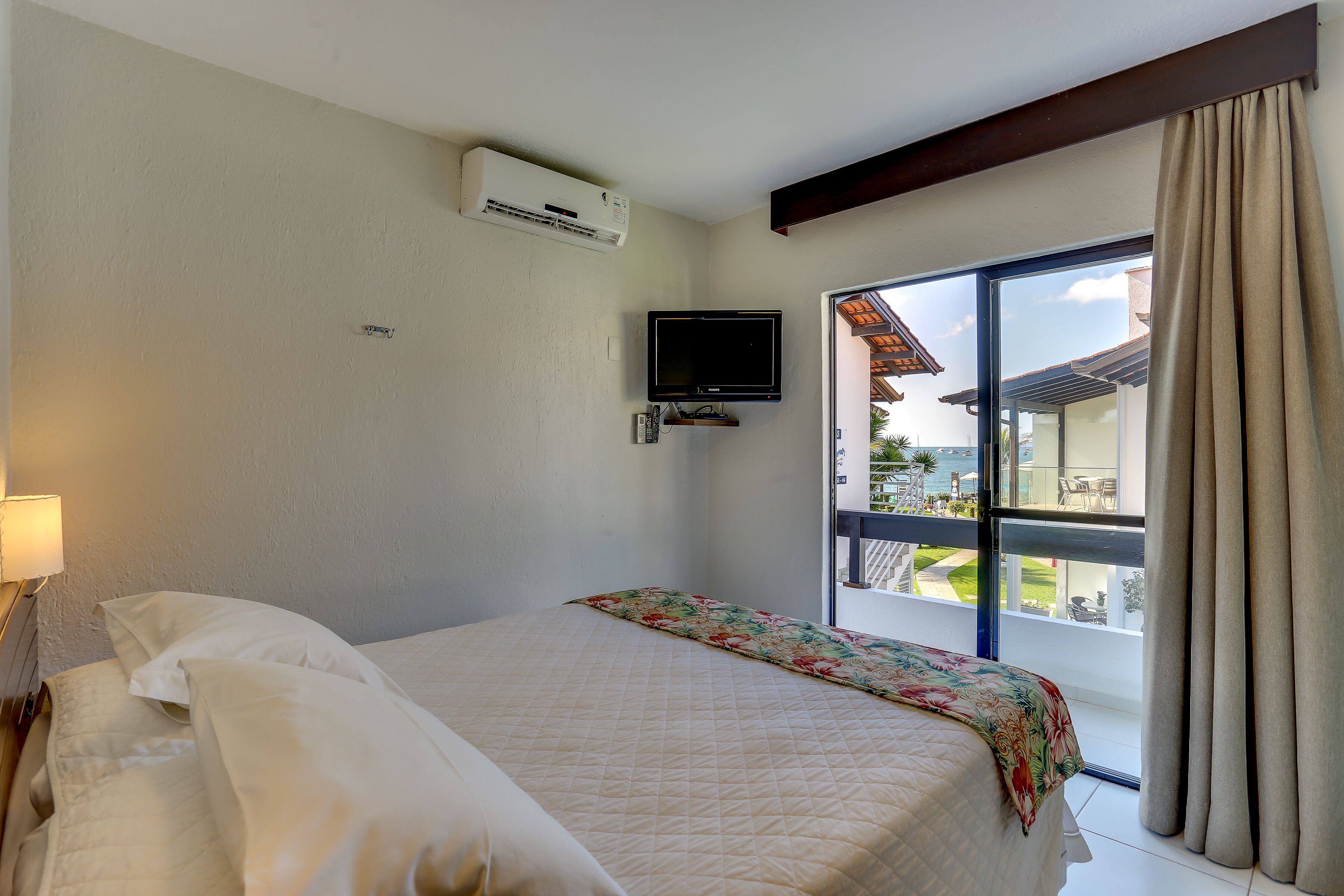 Habitación de hotel con cama, televisión montada en la pared y un amplio ventanal con cortinas.
