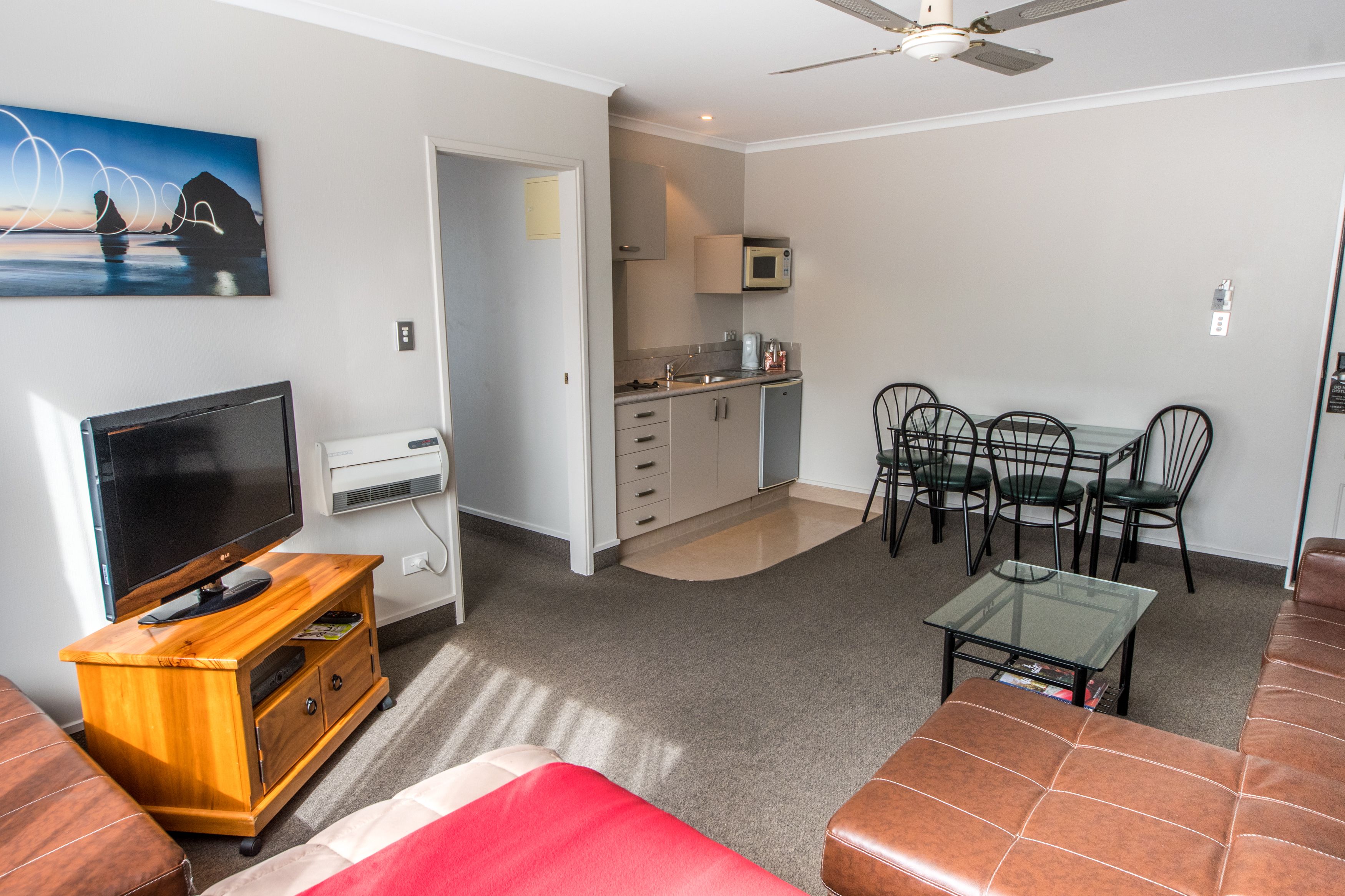 A modern living room with a flat-screen TV, wooden entertainment unit, and a red sofa. There is a dining area with a glass table and chairs, and a kitchen with a microwave and cabinets.