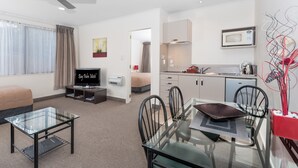 A modern hotel room with a glass dining table, black chairs, a TV, a ceiling fan, a kitchenette, and a bed. - Bay Palm Motel (Tauranga)