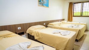 Une chambre d’hôtel avec quatre lits simples, chacun recouvert d’une courtepointe beige et agrémenté d’oreillers. On y trouve une fenêtre avec un rideau, un tableau au mur et un sol carrelé.