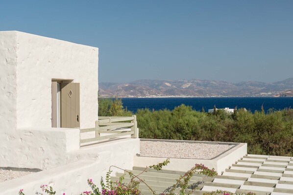 A white building with a door and a bench, overlooking a coastal landscape with mountains and the sea. - Stagones Luxury Villas (Paros)