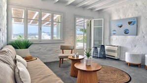 A modern living room with a beige sofa, wooden coffee table, and a view of the countryside through large windows. - Stagones Luxury Villas (Paros)