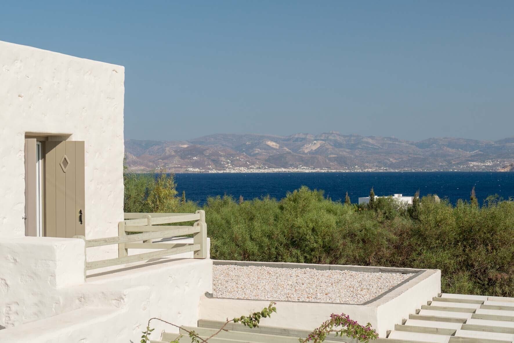 A white building with a door and a wooden bench. There is a view of the sea and distant mountains.