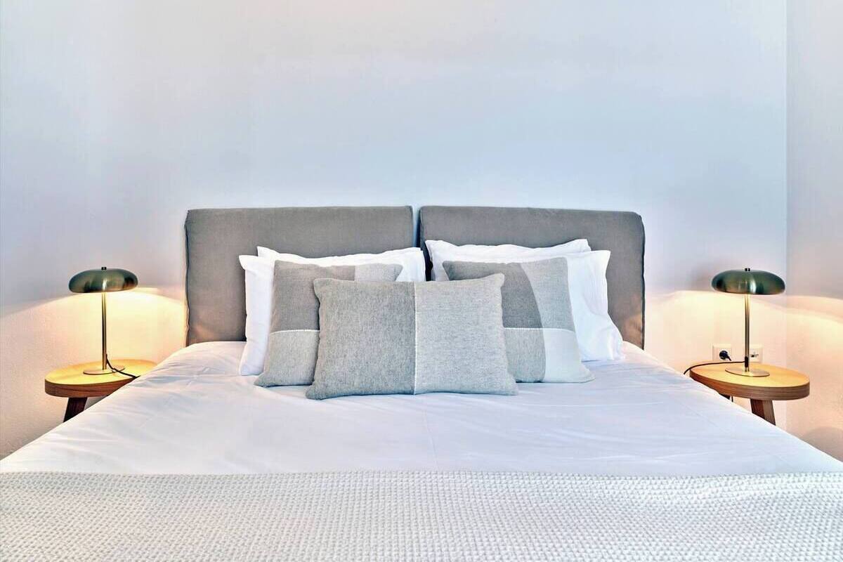 A neatly made bed with a grey and white pillow arrangement, two bedside lamps, and a plain wall in the background.