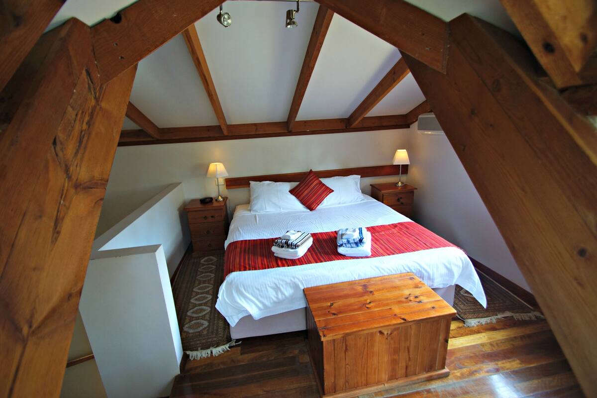 A bedroom with a wooden ceiling, a bed with white bedding and a red pillow, two bedside tables with lamps, and a wooden bench.