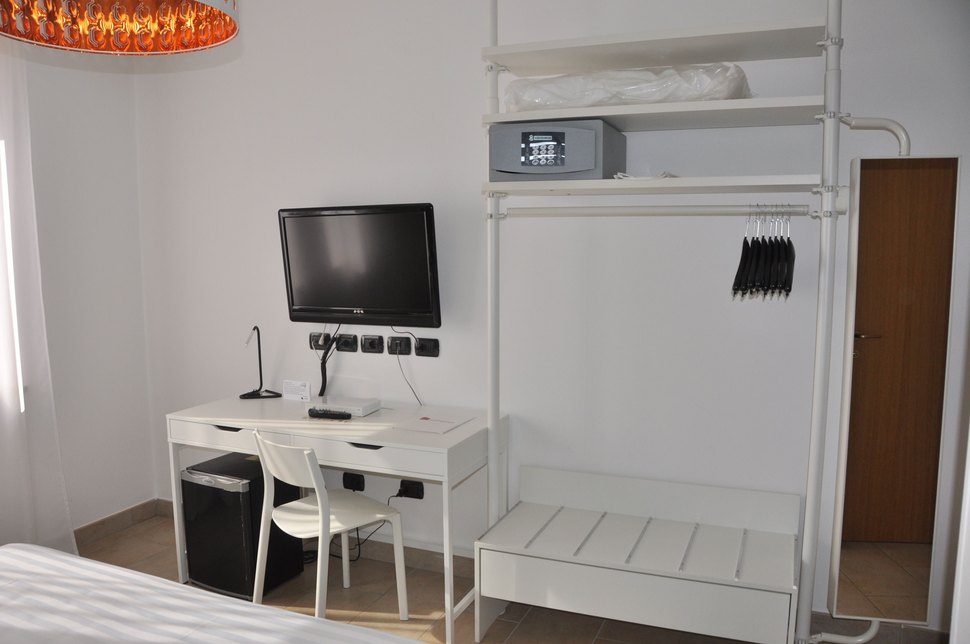 Une pièce de taille modeste, avec un bureau blanc, un téléviseur noir, une chaise blanche, une petite poubelle et un lit superposé avec une étagère au-dessus.