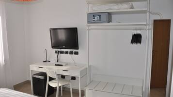 Une pièce de taille modeste, avec un bureau blanc, un téléviseur noir, une chaise blanche, une petite poubelle et un lit superposé avec une étagère au-dessus.