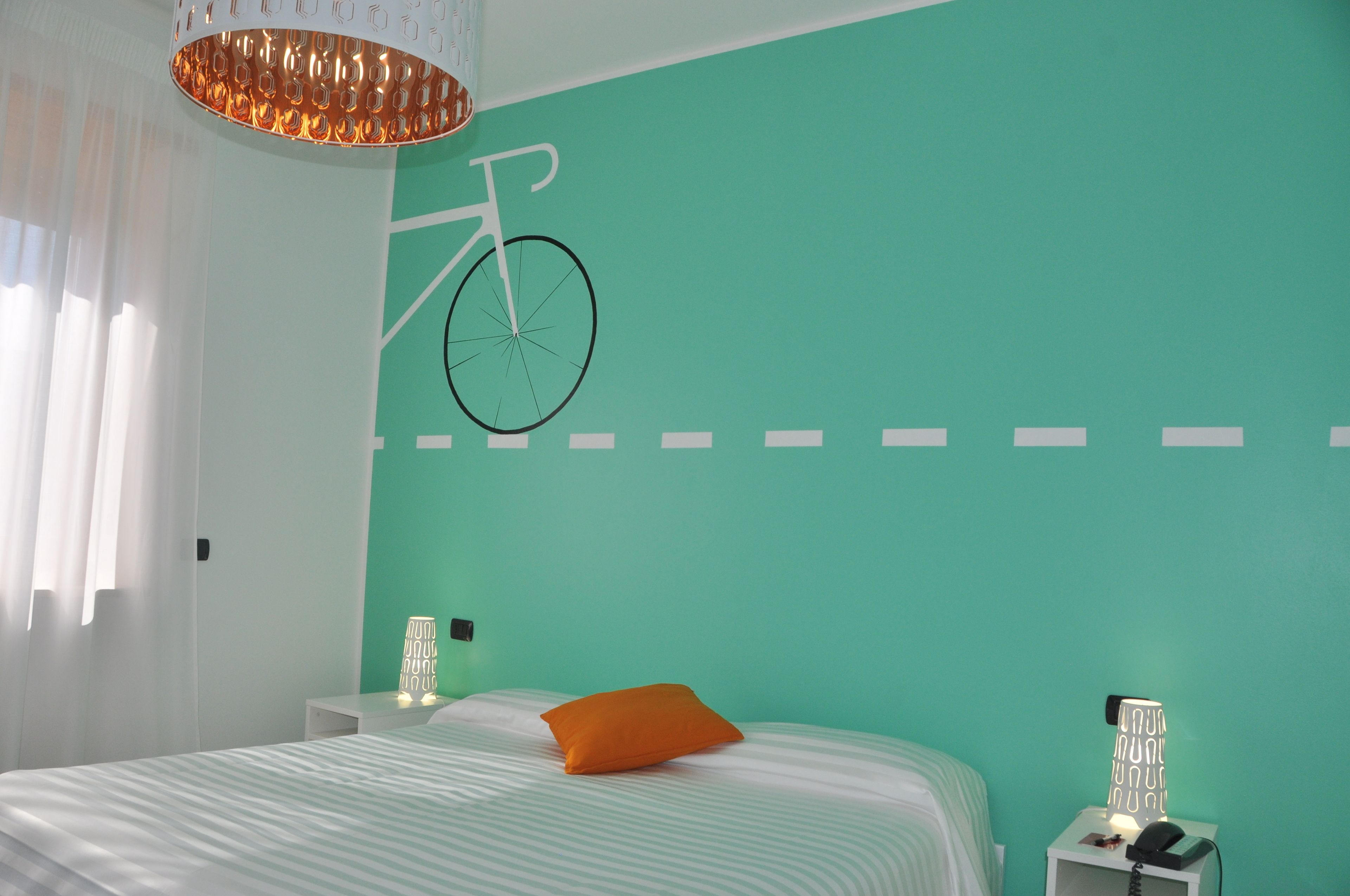 A bedroom with a teal wall, a bed with striped bedding, an orange pillow, a nightstand with a lamp, and a wall-mounted bicycle decoration.