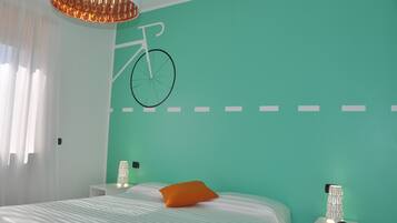 A bedroom with a teal wall, a bed with striped bedding, an orange pillow, a nightstand with a lamp, and a wall-mounted bicycle decoration.
