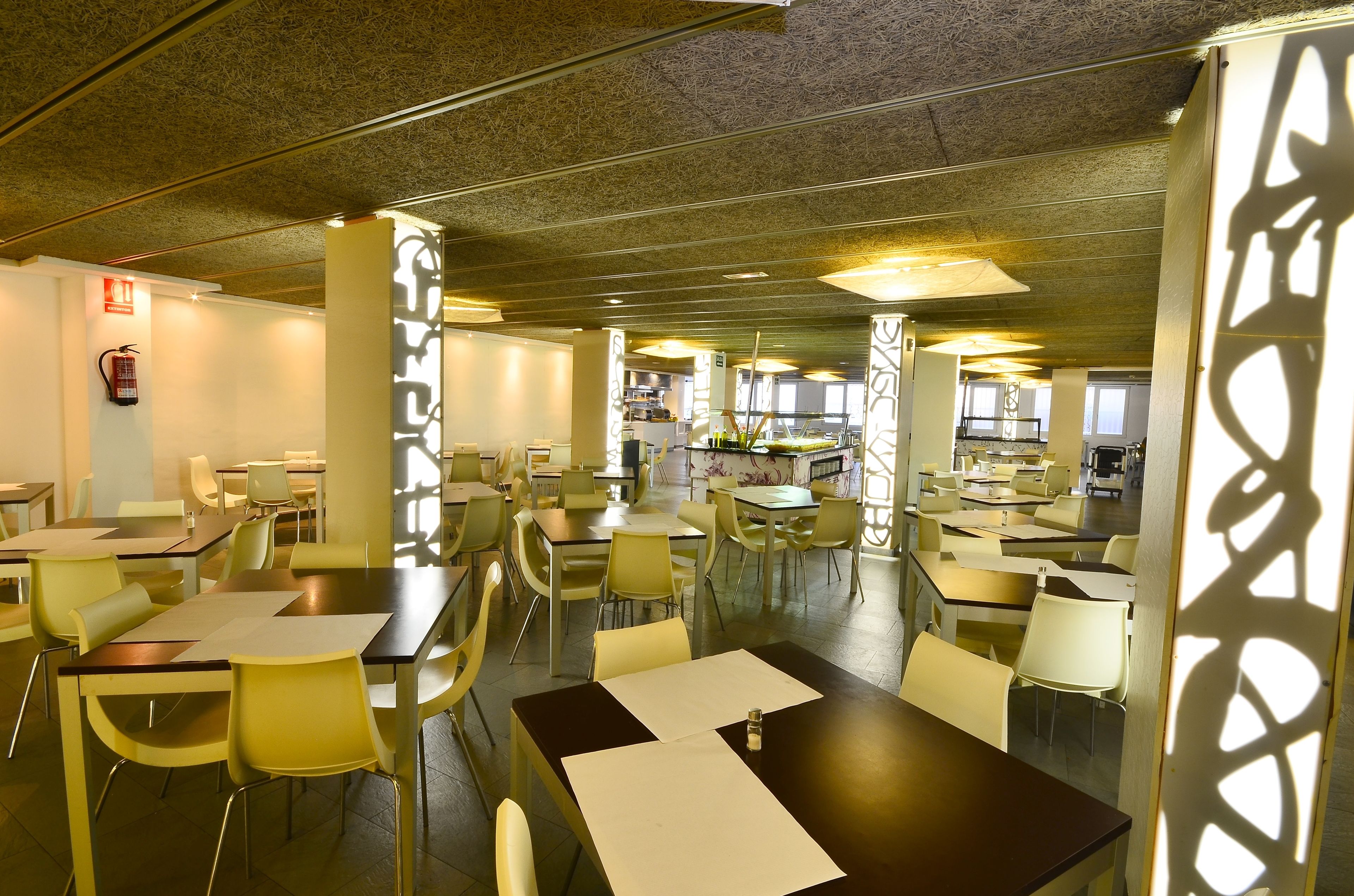 Una cafetería amplia con varias mesas y sillas, un extintor en la pared y vista al área de la cocina de fondo.