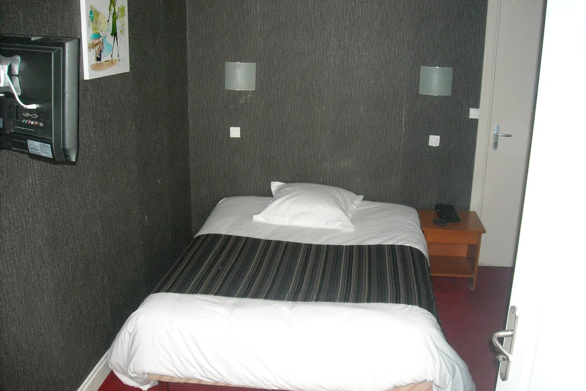 Habitación de hotel con una cama individual, una mesita de noche, un televisor montado en la pared y dos apliques.