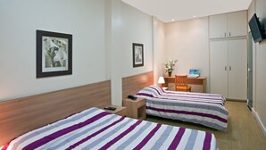 Quarto de hotel com duas camas, uma escrivaninha, uma cadeira, uma televisão e um guarda-roupa.