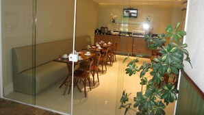 Uma sala de jantar com uma mesa comprida posta para refeição, uma cozinha com televisão e uma divisória de vidro.