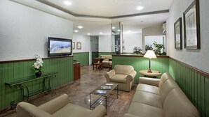 Um hall com paredes revestidas de verde, sofás bege, uma mesa de centro, uma televisão e um balcão de recepção.