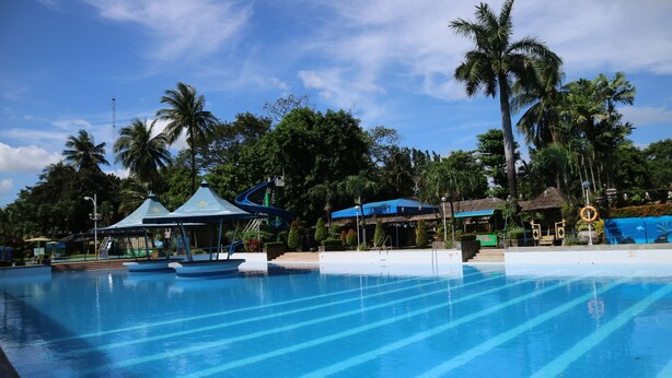 A large outdoor swimming pool with clear blue water, surrounded by palm trees and shaded areas with blue canopies. - Riverview Resort & Conference Center (Calamba)