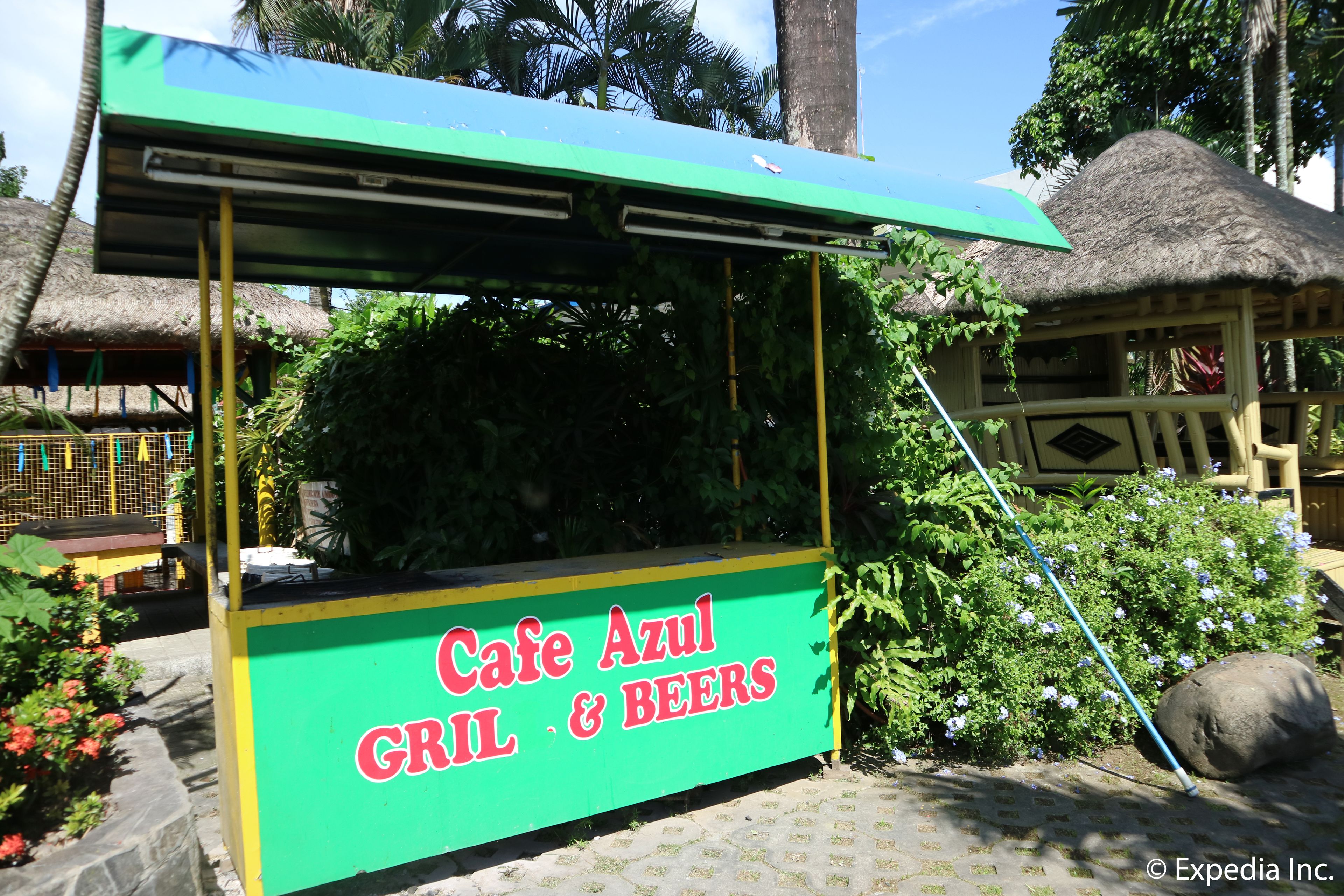 Um quiosque de comida chamado Cafe Azul Grill & Beers, com um letreiro verde e amarelo, cercado por plantas tropicais e uma estrutura com telhado de palha.