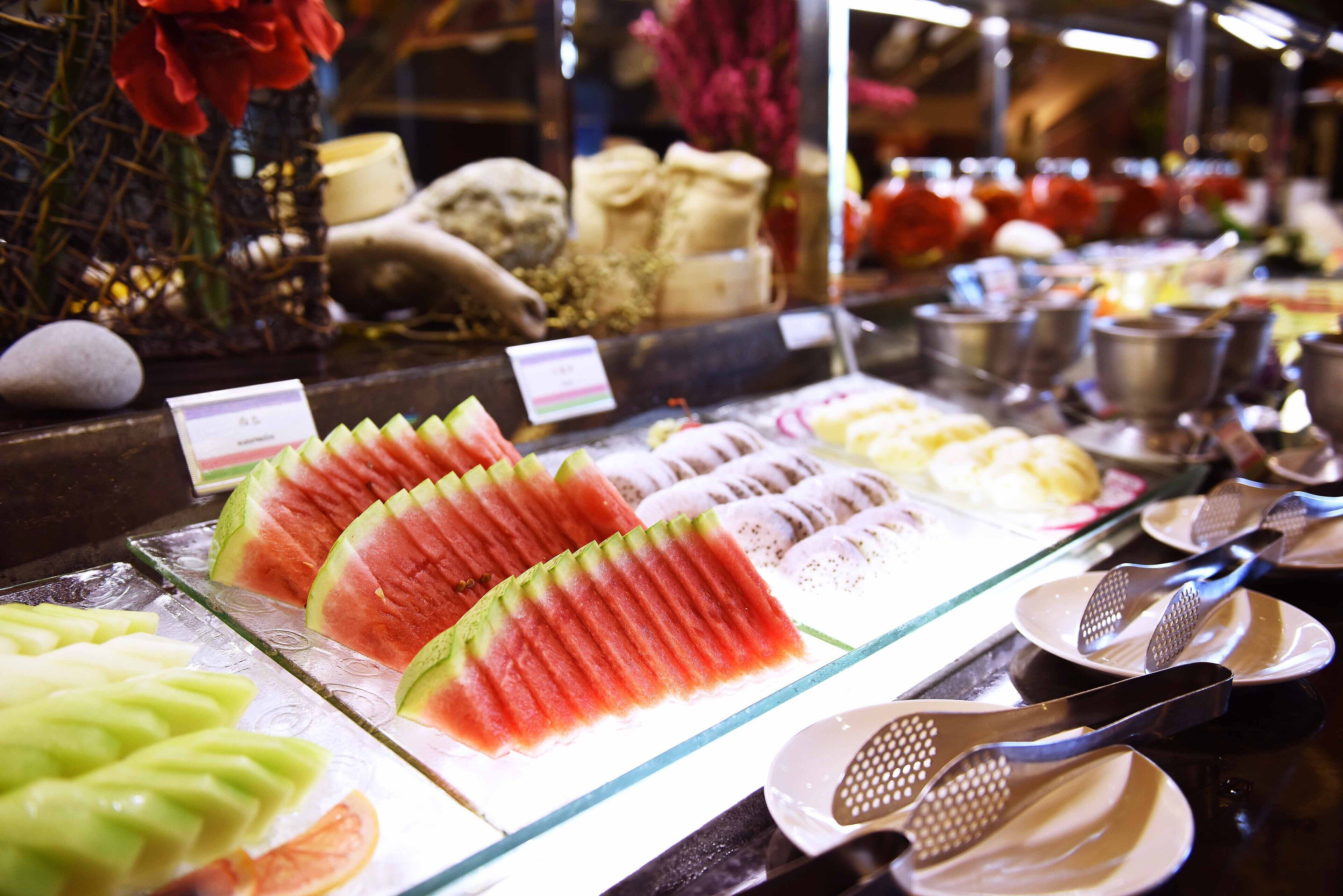 A display of various fruits and vegetables, including watermelon and green apples, arranged on a glass counter.