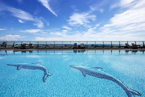 Una piscina con mosaicos de delfines, sillas de descanso y vistas de la ciudad.