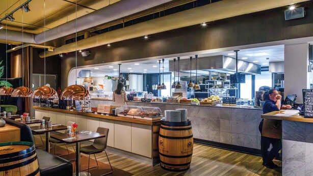 A modern restaurant with a bar area, wooden barrels, and a visible kitchen.